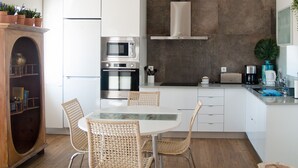 Cuina moderna amb armaris blancs, forn encastrat, microones i una campana extractora d'acer inoxidable. Hi ha una taula de dinar amb cadires de vímet i un moble de fusta amb plantes.
