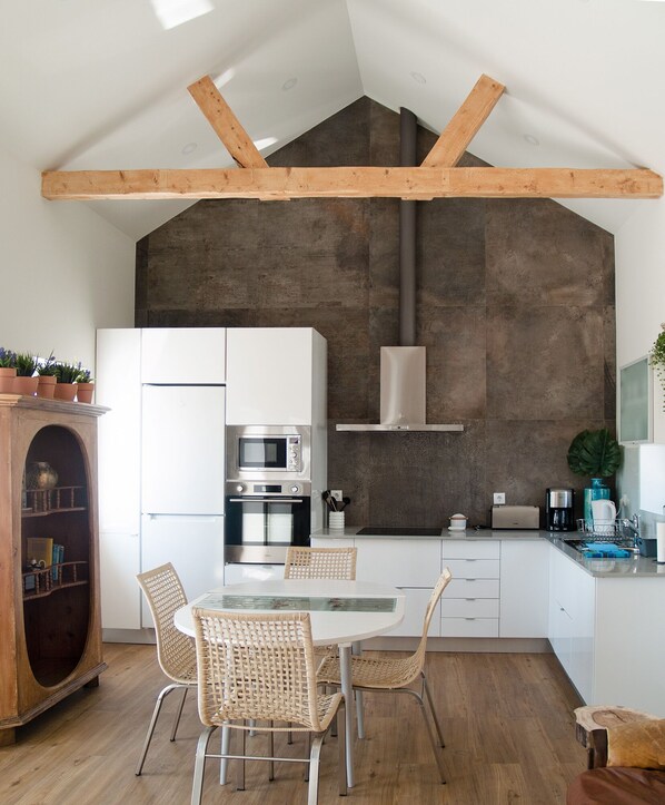 A modern kitchen with white cabinetry, a built-in oven, microwave, and a stainless steel range hood. There is a dining table with wicker chairs and a wooden cabinet with plants.