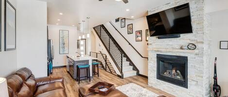 A modern living room with leather sofas, a fireplace, a TV mounted on the wall, and a staircase leading to the upper floor.