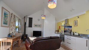 A cozy living room with a brown leather sofa, wooden dining table, and a TV. A kitchen with white cabinets and a stove is visible. - Wonderful Wallow barn - Close  to Sennen Cove  & Porthcurno Cove & Coastal Path (Penzance)