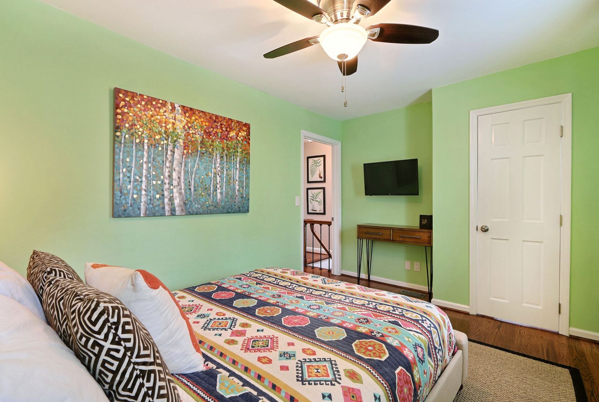 A bedroom with a bed, a ceiling fan, a painting, a TV, and a door.