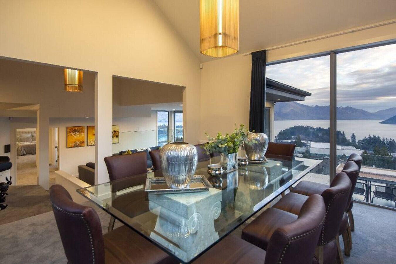 A modern dining area with a glass table, leather chairs, and a view of a lake and mountains.