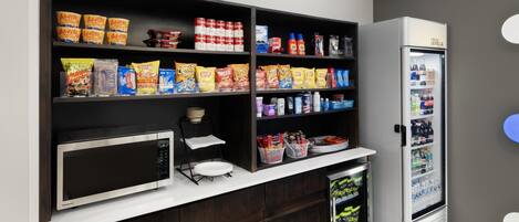 A kitchenette with a microwave, shelves stocked with snacks, a refrigerator, and a coffee machine.