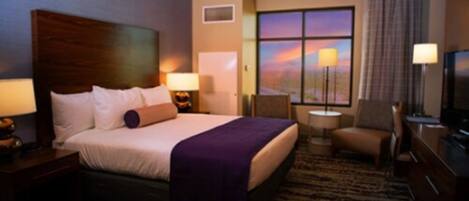 A hotel room with a large bed, two bedside tables with lamps, a small round table, a chair, a TV, and a window with a sunset view.