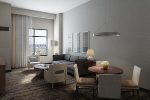 Une chambre d’hôtel avec un canapé, deux fauteuils, une petite table, une lampe et une vue sur la ville, visible à travers une grande fenêtre.