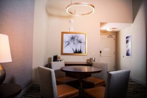 Habitación de hotel con una mesa de comedor redonda, cuatro sillas tapizadas, una obra de arte en la pared, una puerta y una pequeña mesita de noche con una lámpara.