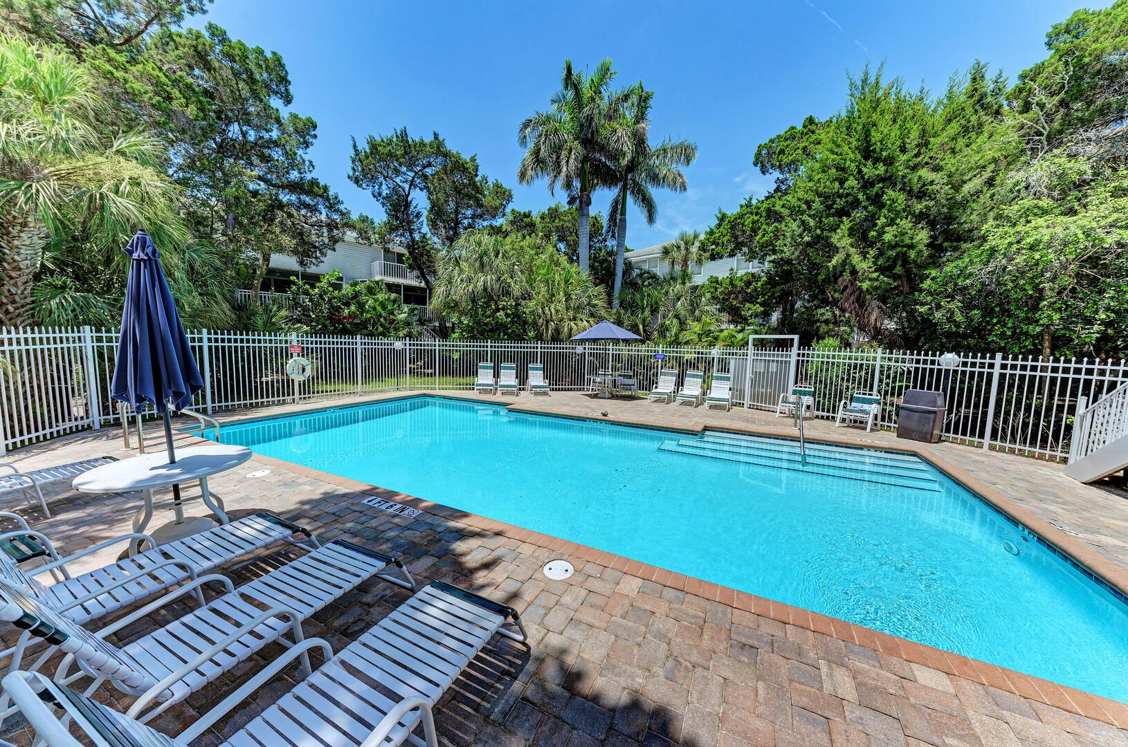 A rectangular swimming pool with clear blue water, surrounded by a white fence. There are lounge chairs and umbrellas along the poolside.