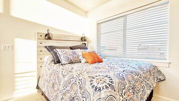 Un dormitorio con cama, cabecera, ventana con persianas y un interruptor de luz en la pared.