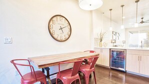 Un coin repas avec une table en bois et quatre chaises rouges, une horloge murale, des luminaires suspendus et un espace cuisine équipé d’un lave-vaisselle.