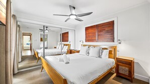 Une chambre d’hôtel avec un grand lit, un ventilateur de plafond, une tête de lit en bois, une table de chevet, un miroir et une fenêtre avec des volets en bois.