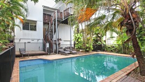 Une piscine avec une terrasse en bois, deux chaises longues et un escalier menant à un balcon.