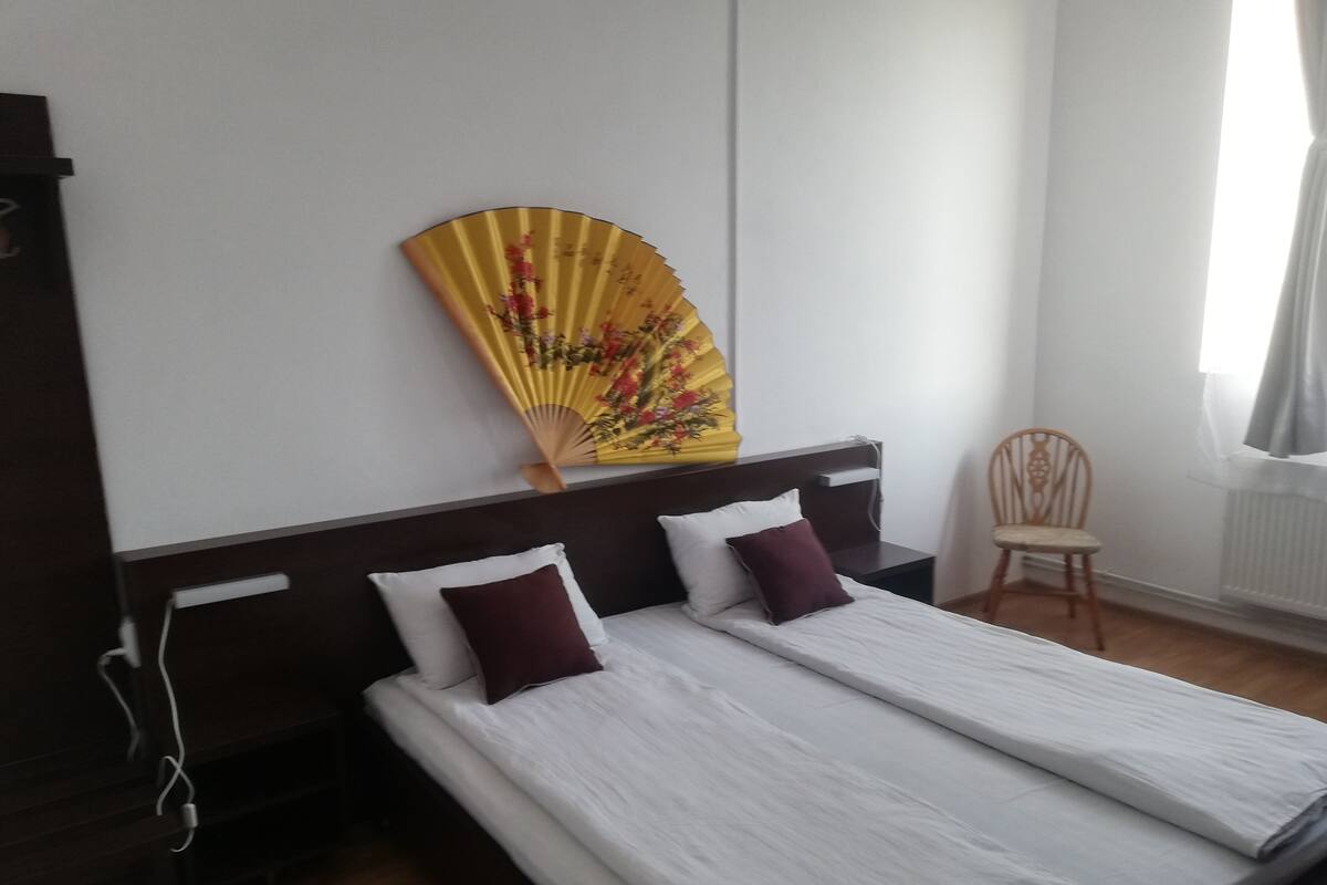 Une chambre d’hôtel avec deux lits simples, une table de chevet en bois, une chaise et un éventail décoratif jaune.