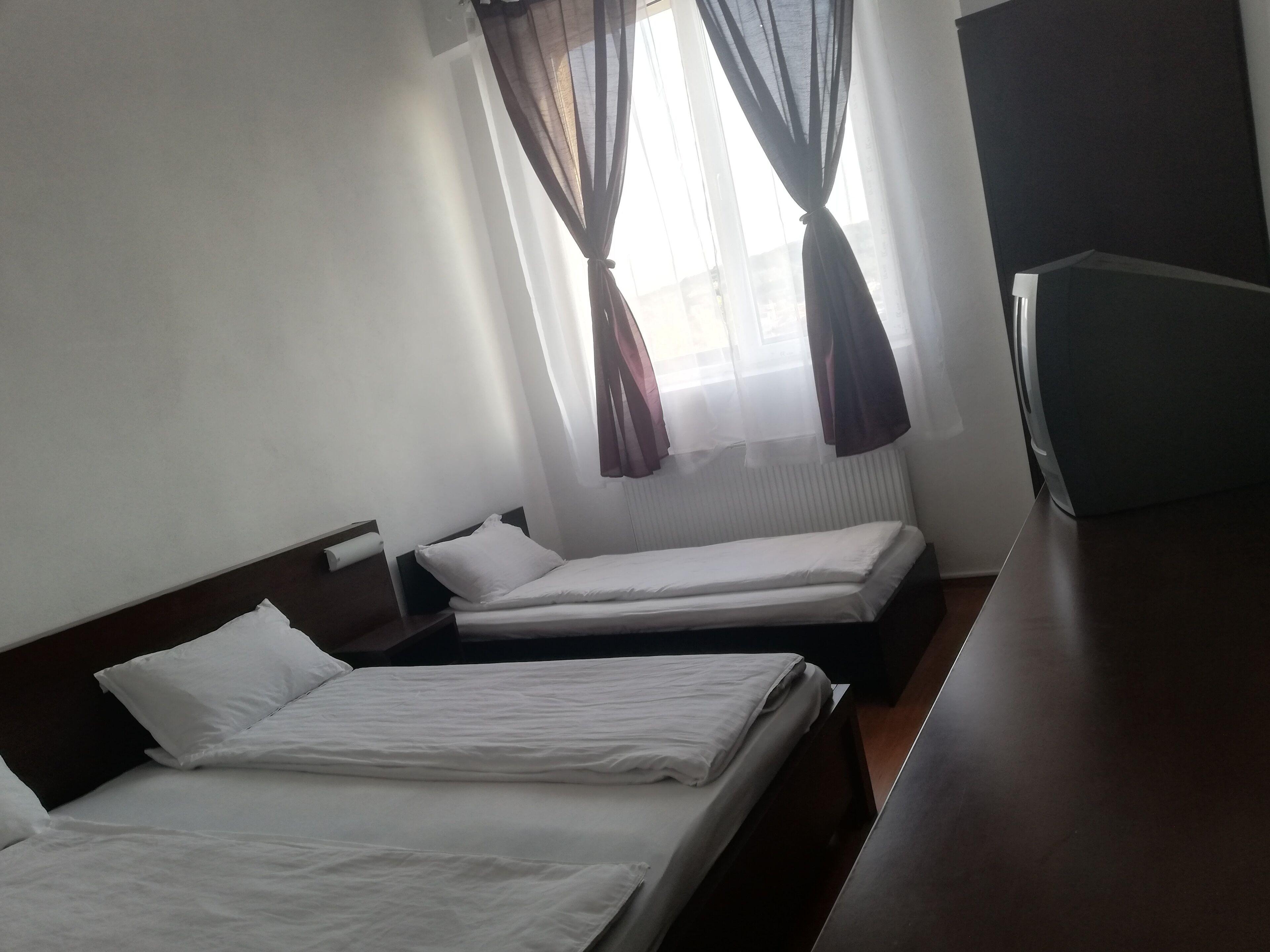 A hotel room with two single beds, a wooden headboard, a small bedside table, a TV, and sheer curtains.
