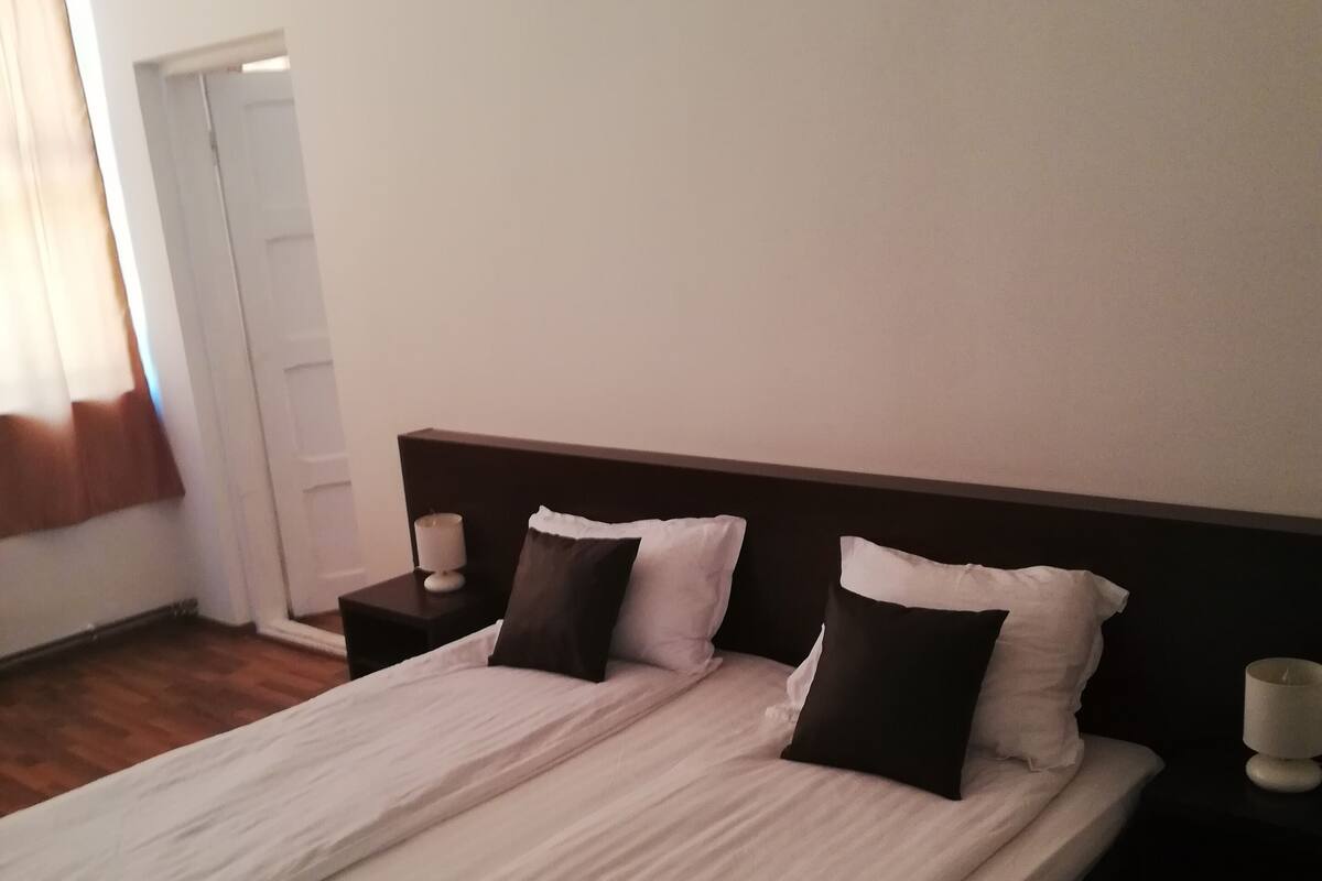 Un lit bien fait, avec une couette rayée blanche et grise, deux oreillers noirs et une tête de lit en bois.
