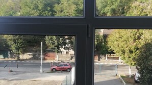 Une vue à travers une fenêtre sur un parking où l’on aperçoit une voiture rouge, une clôture et, au loin, une colline parsemée d’arbres.