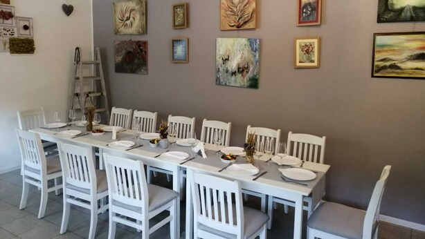 Un espace repas avec une longue table dressée pour six personnes, entourée de chaises blanches. Les murs sont ornés de divers tableaux encadrés.
