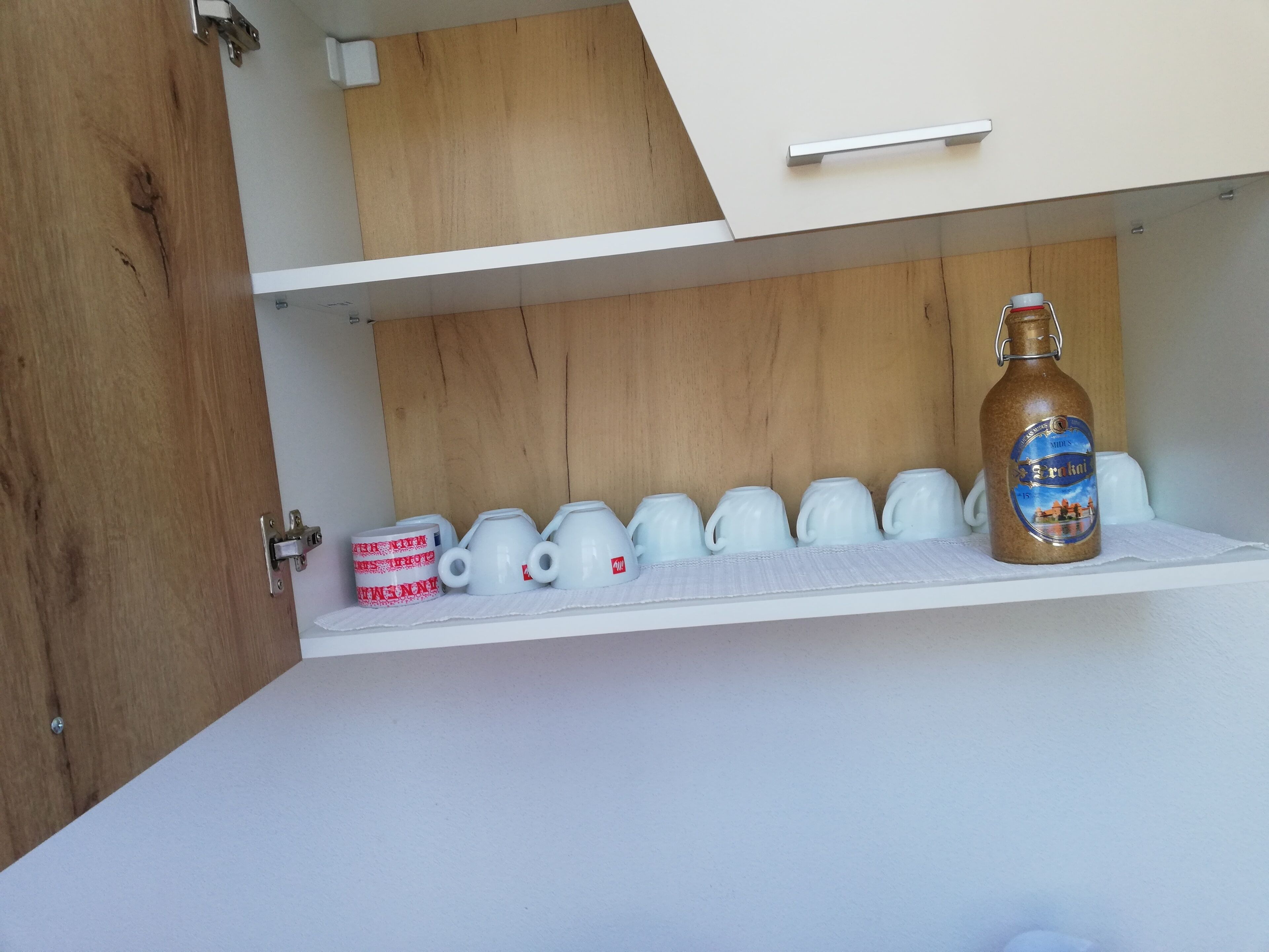 A kitchen with white cabinets, a wooden door, and a shelf with white mugs and a bottle.