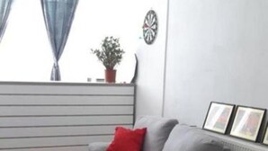 Un salon avec un canapé gris, une petite table d’appoint, un coussin rouge, un jeu de fléchettes et une plante en pot.