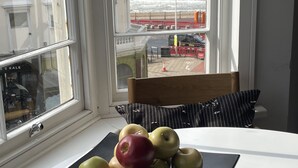 A table with a fruit bowl containing apples, a chair, and a view of the beach and ocean through a window.