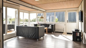 A spacious room with a wooden ceiling, large windows, a black sofa, and a television.
