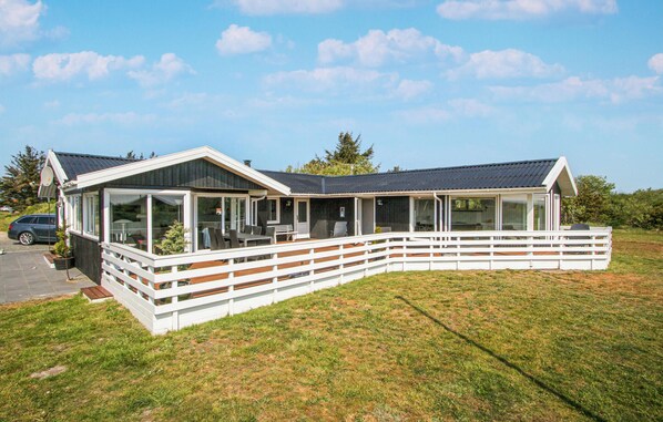 Et moderne hus i to etager med stor terrasse og glasdøre, omgivet af et hvidt hegn.