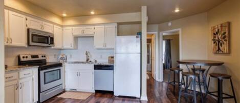 A kitchen with white cabinets, a microwave, stove, and refrigerator. There are wooden floors and a bar area with stools.