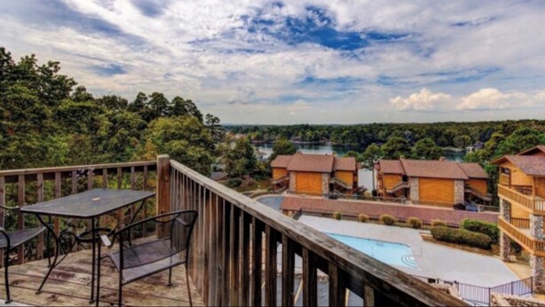 Une terrasse en bois avec des meubles d’extérieur, une piscine et une vue sur un lac entouré d’arbres et de bâtiments.