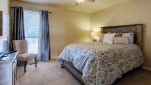 Une chambre à coucher comprenant un lit, un bureau, une chaise, un ventilateur de plafond et une fenêtre avec des rideaux.