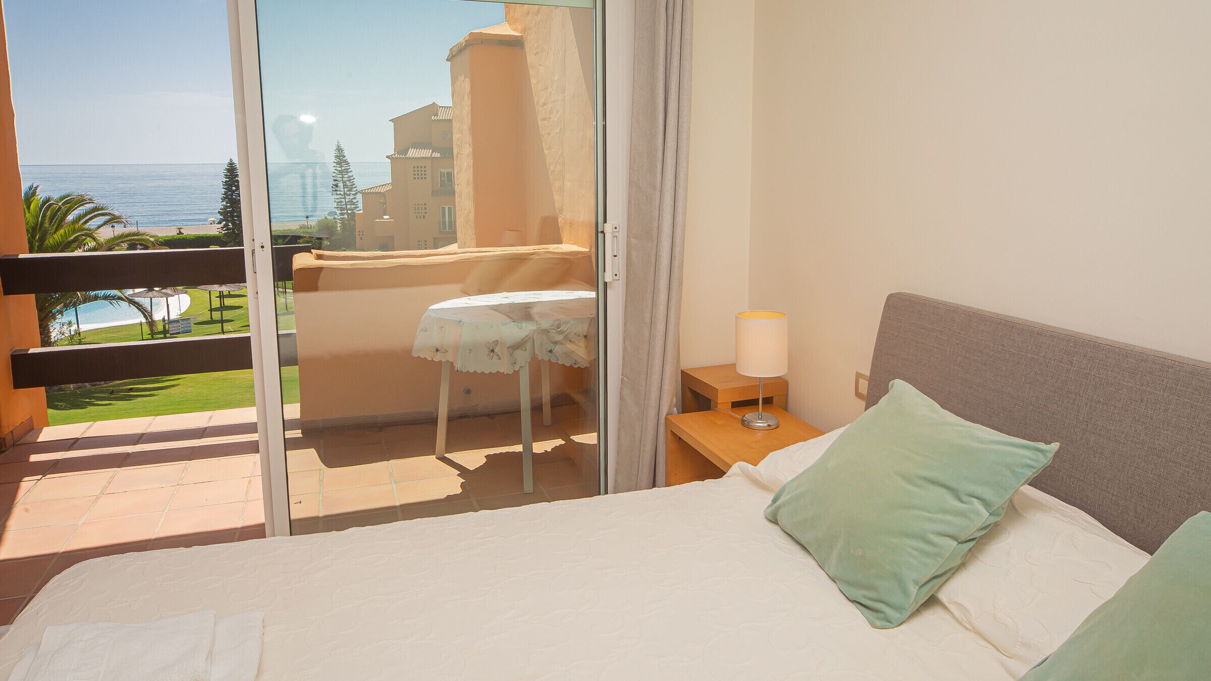 Une chambre avec un lit, une table de chevet, une table recouverte d’une nappe, un balcon avec une table, et une vue sur la mer et les bâtiments.
