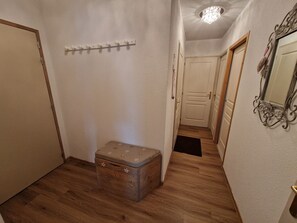 Um corredor com piso de madeira, um pequeno baú, um porta-roupas e um espelho na parede.
