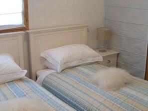 Quarto com duas camas, cada uma com roupa de cama listrada e almofadas brancas. Tem uma mesa de cabeceira com abajur e uma gavetinha.