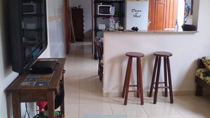 Ein Wohnzimmer mit Fernseher, Holztisch und einer Zierpflanze. Im Hintergrund ist eine Küche mit Mikrowelle, Kühlschrank und Holzschränken zu sehen.