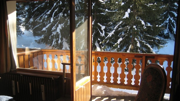 A room with a view of snow-covered trees through a window, a wooden deck outside, and a patterned rug inside.