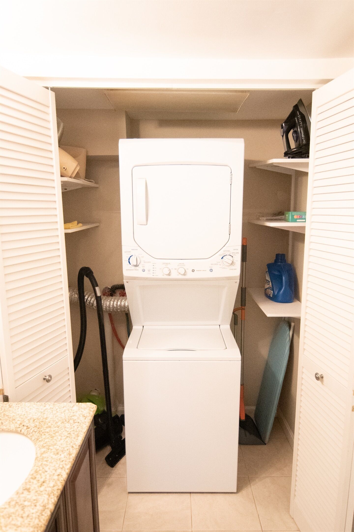 A compact laundry room with a front-loading washer and dryer, a shelf with cleaning supplies, and a vent hose.