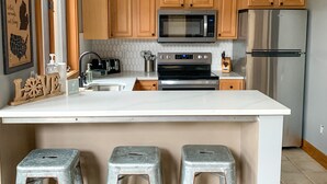 Una cocina moderna con armarios de madera, un refrigerador de acero inoxidable, un microondas y un horno empotrado. Hay tres taburetes de metal en la isla.