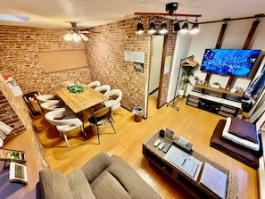 A modern living room with a brick wall, a wooden dining table, white chairs, a sofa, a TV, and a small plant. - Hotel El Shinjuku 2 (Tokyo)