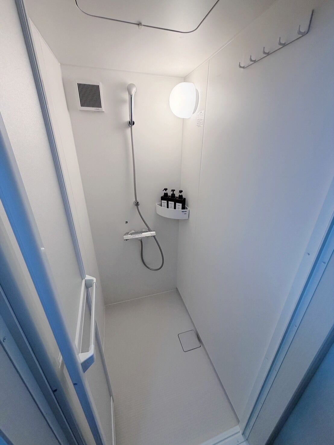 A small, white, enclosed shower area with a handheld showerhead, a wall-mounted soap dispenser, and a small mirror.