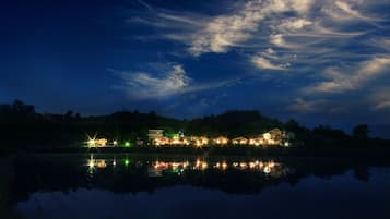 A lakeside hotel with multiple illuminated buildings reflecting on the water, surrounded by hills.