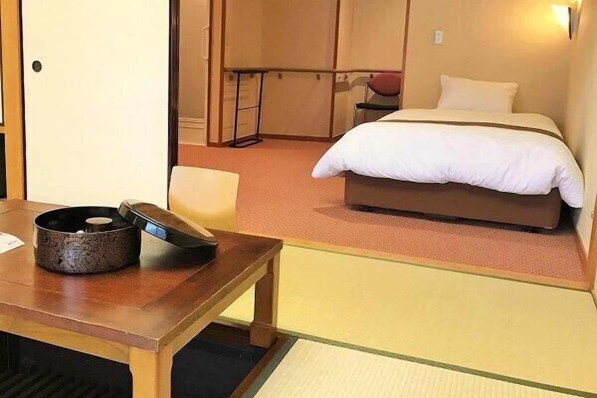 Habitación de hotel con cama, una mesa de madera con un cuenco decorativo, una silla y una mesita con un objeto rojo.