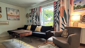 A living room with a ceiling fan, a sofa, a coffee table, and a side table. There is a window with curtains, a rug, and a painting on the wall. - Sea Grape Escape Mid-Century Modern Pool home! (North Palm Beach)
