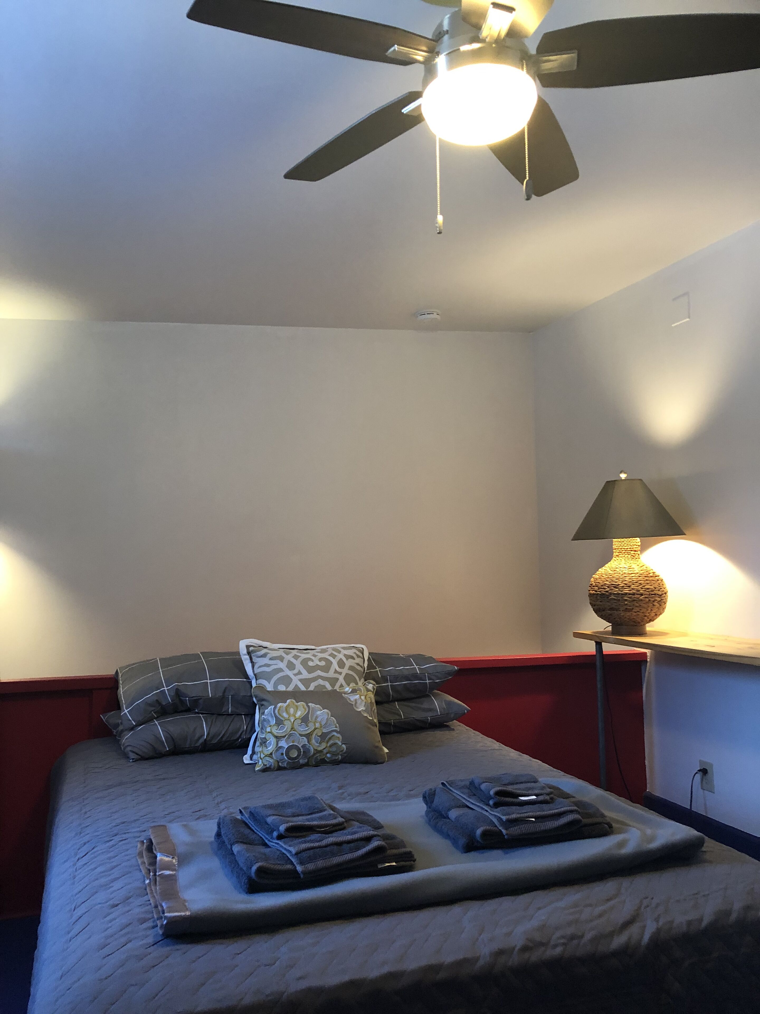 A neatly made bed with pillows and folded towels, a ceiling fan, and a lamp on a nightstand.
