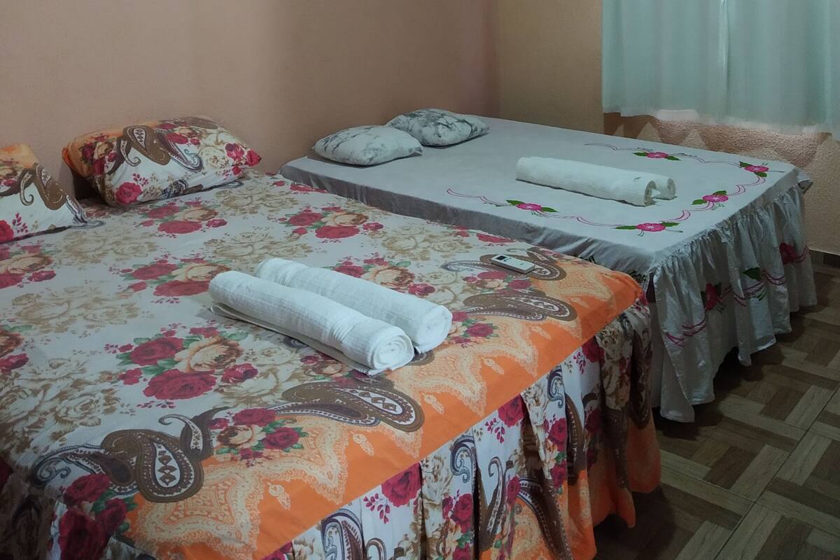 A hotel room with two beds, each with floral-patterned bedding and pillows. There is a ceiling fan, an air conditioning unit, and a window with curtains.