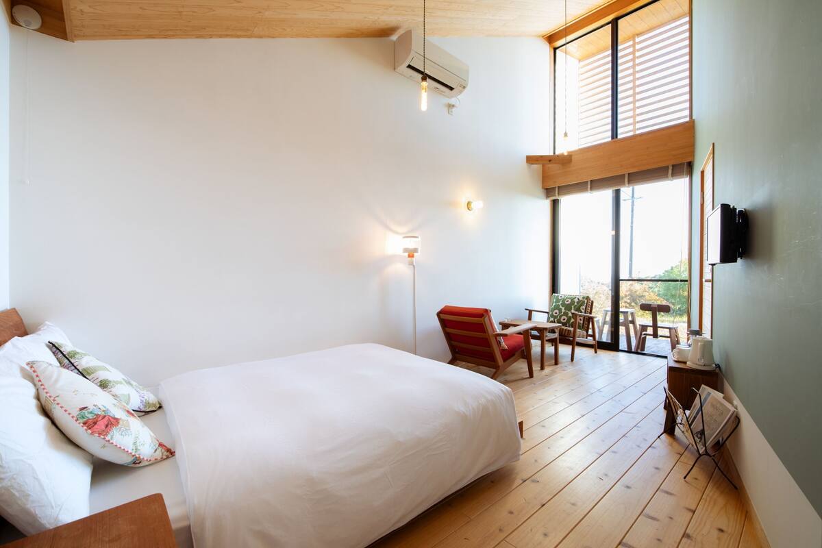 A bedroom with a large bed, a chair, a table, and a view of the outdoors through a window.