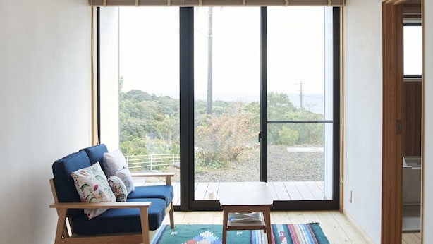 A bedroom with a large bed, a blue sofa, a small table, and a view through a large window. - Ananda Chillage (Yakushima)