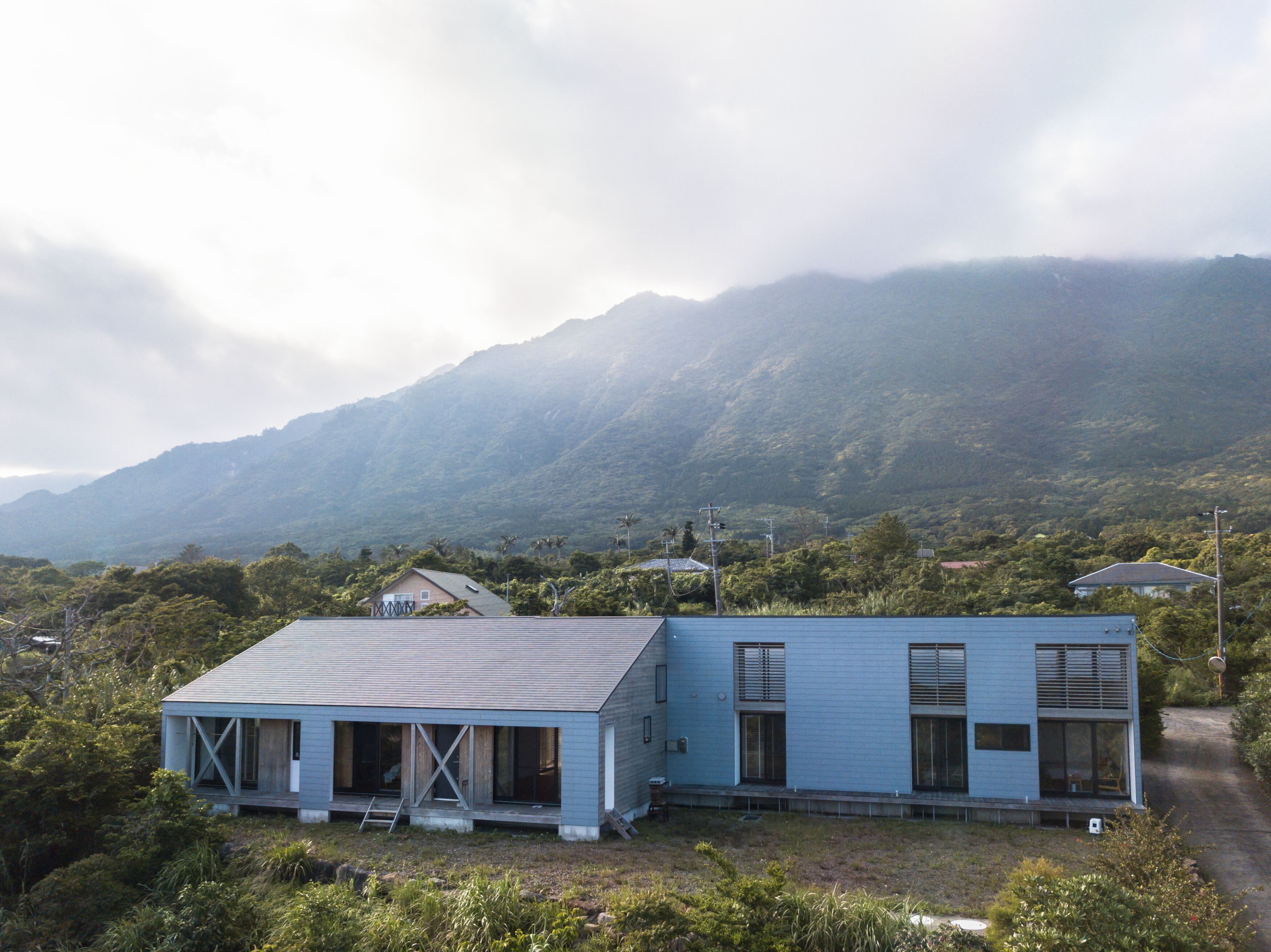 A modern two-story house with a flat roof and large windows, surrounded by lush greenery and mountains in the background.