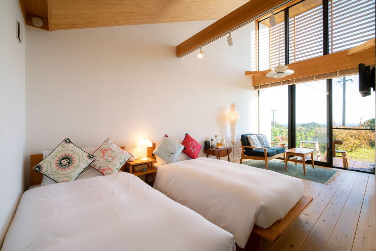 A bedroom with two beds, a nightstand, a lamp, and a view of the outdoors.
