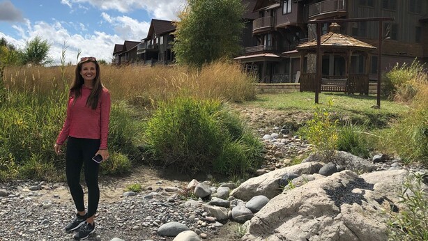 A person standing on a rocky path with large rocks and sparse vegetation. In the background, there are wooden houses with balconies and a clear sky with clouds.