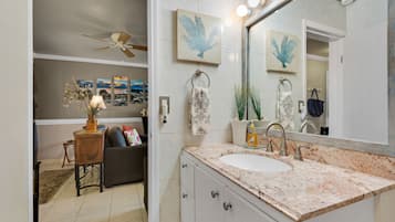 A bathroom with a white sink, marble countertop, and a large mirror. There are two wall-mounted lights above the mirror and a ceiling fan in the adjacent room.