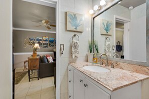 Une salle de bain avec un lavabo blanc, un comptoir en marbre et un grand miroir. Deux appliques murales sont fixées au-dessus du miroir, et un ventilateur de plafond se trouve dans la pièce adjacente.