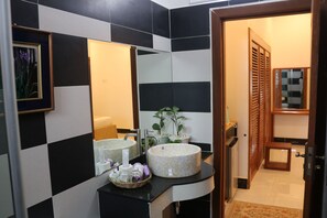 A bathroom with a black and white checkered wall, a large mirror, two sinks, a basket of toiletries, and a wooden door leading to another room.
